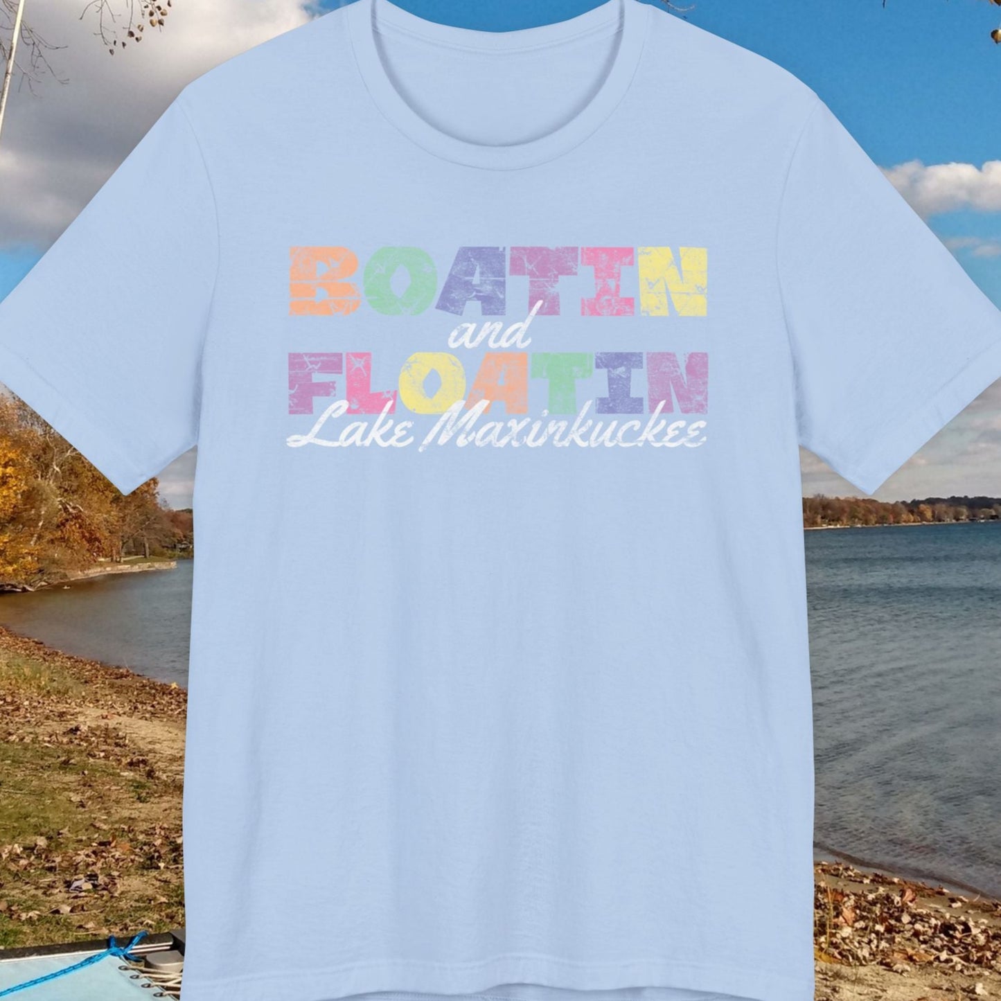 Light blue t-shirt with colorful BOATIN AND FLOATIN LAKE MAXINKUCKEE text on a lake shore background