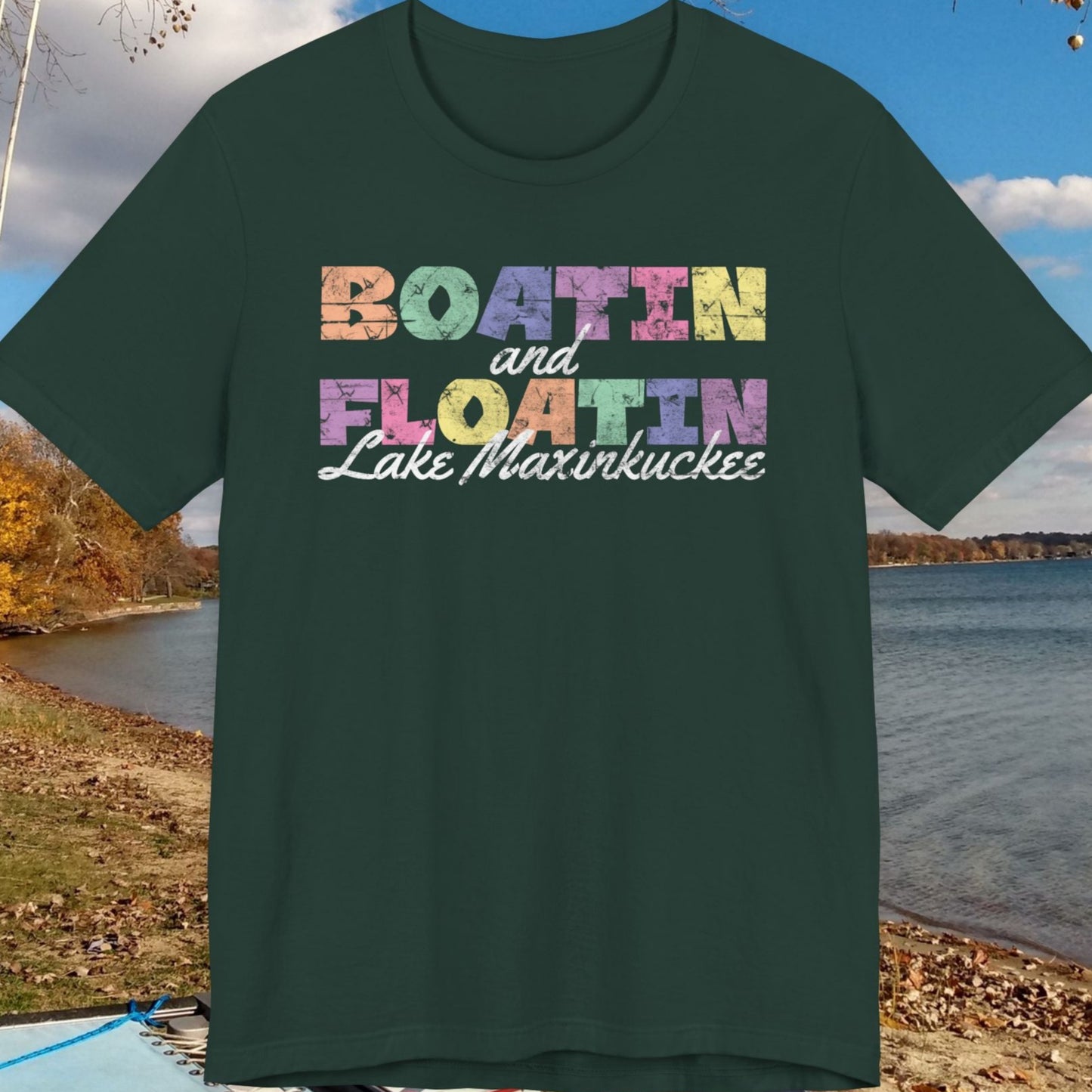 Green t-shirt with colorful text 'Boatin and Floatin Lake Maxinkuckee' against a lakeside background