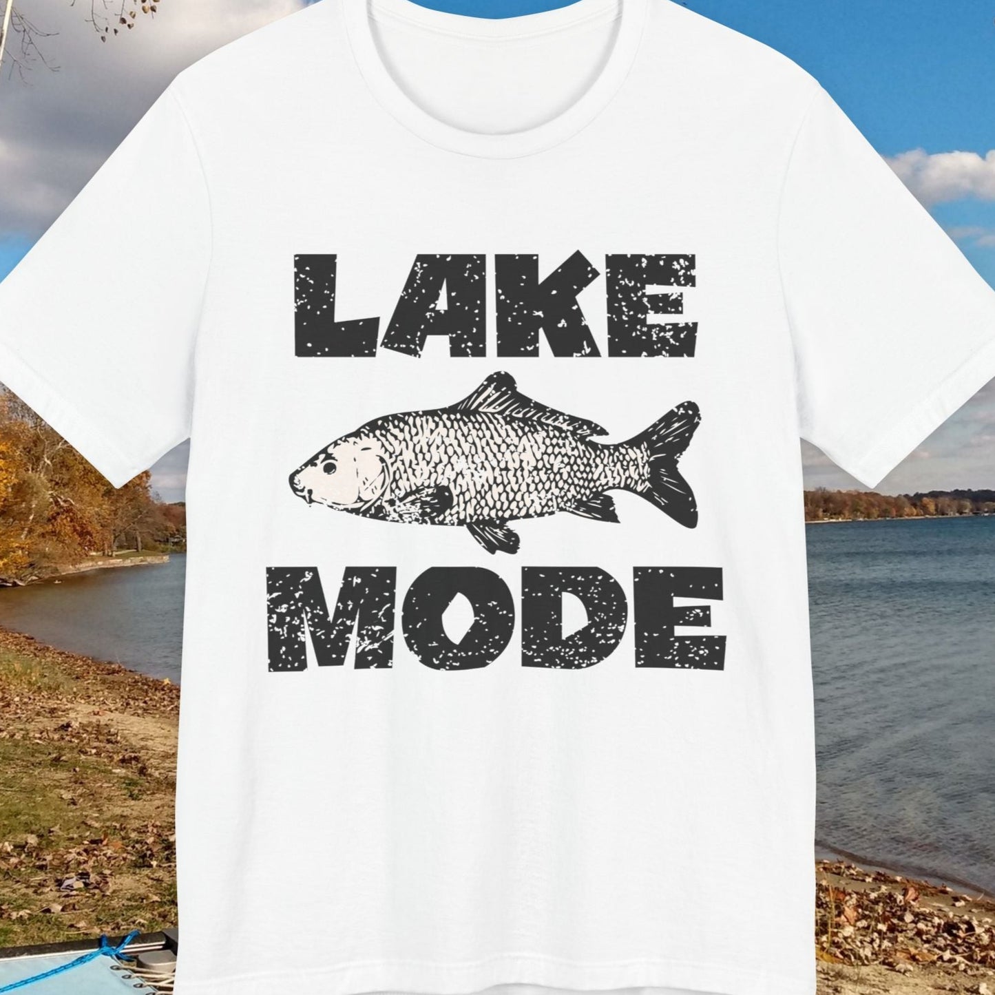 White t-shirt with 'Lake Mode' graphic featuring a fish, displayed outdoors by a lake.
