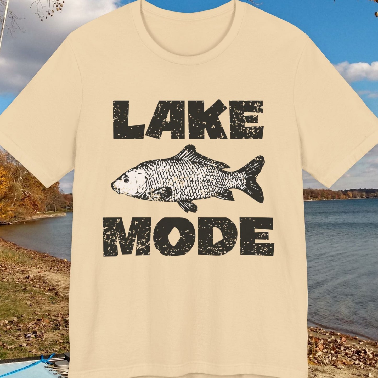 Beige t-shirt with 'Lake Mode' graphic featuring a fish, displayed outdoors by a lake.