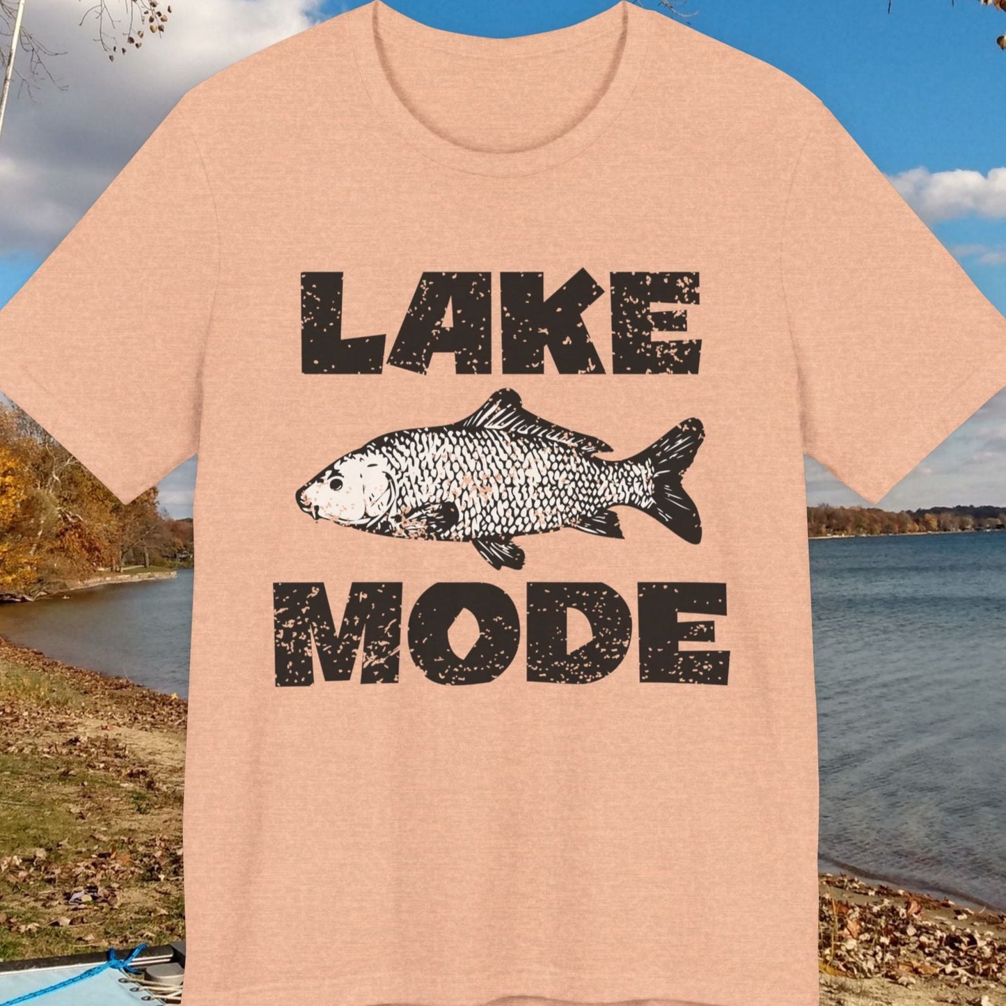 Beige t-shirt with 'Lake Mode' graphic featuring a fish, placed outdoors by a lake.