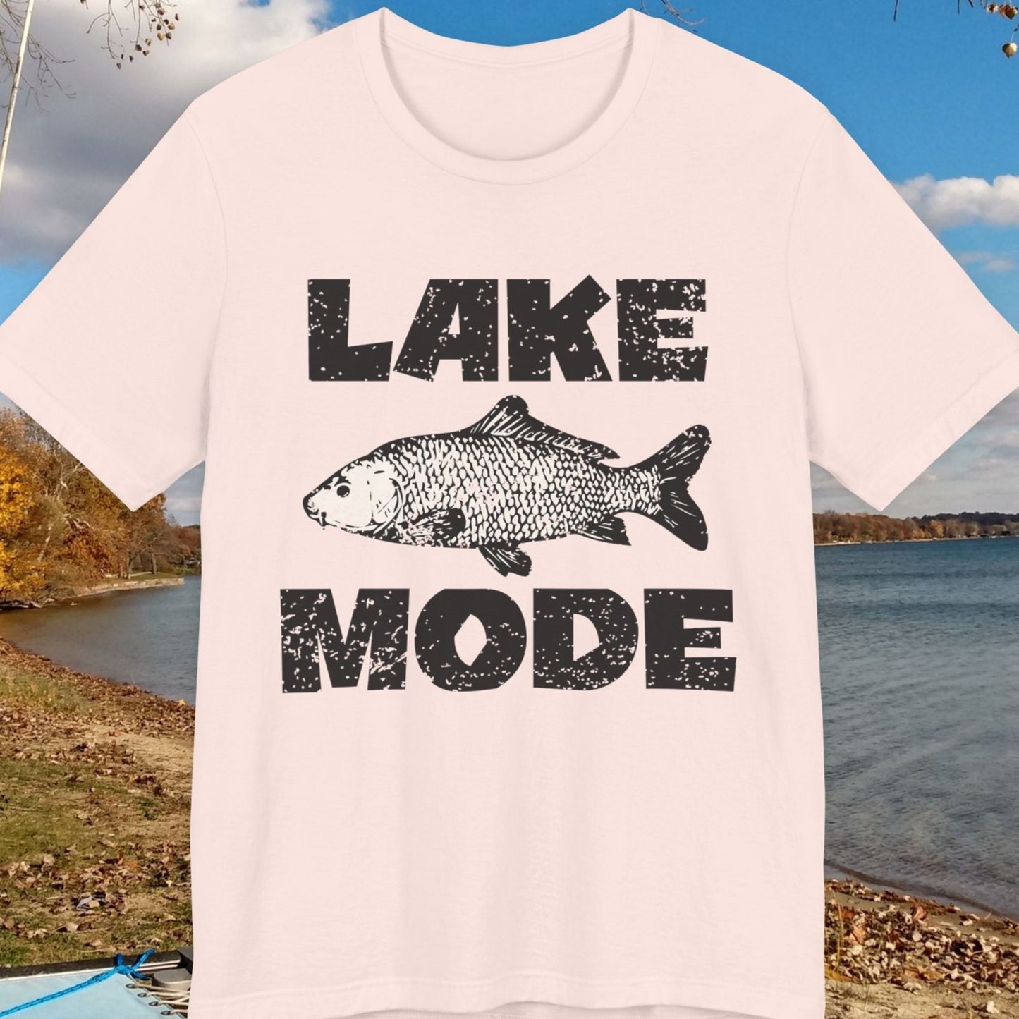 T-shirt with 'Lake Mode' design featuring a fish, placed outdoors by a lake.