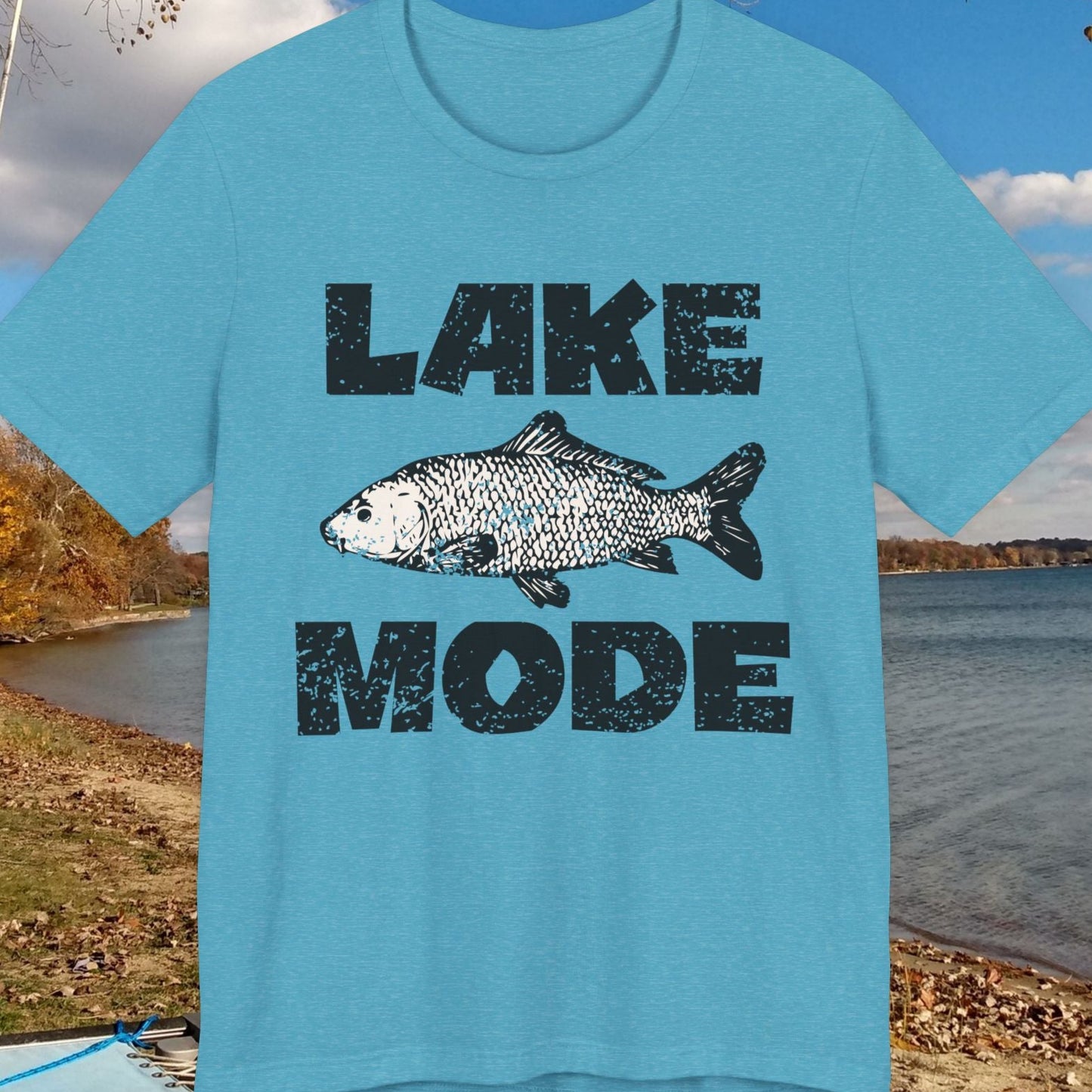 Blue t-shirt with 'Lake Mode' design featuring a fish, placed outdoors by a lake.