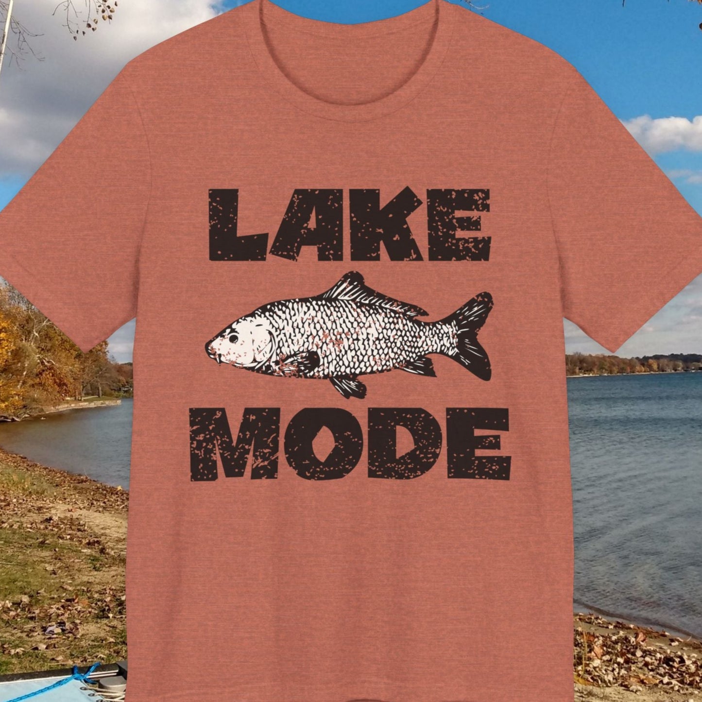 T-shirt with 'Lake Mode' design featuring a fish, displayed outdoors by a lake.