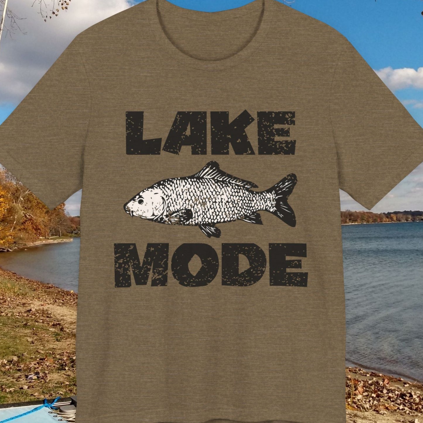 T-shirt with 'Lake Mode' design featuring a fish, placed outdoors by a lake.